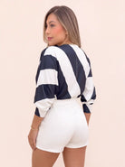 Vista posterior de blusa estampada azul y blanca para mujer, con líneas anchas, mangas amplias y puños ajustados, combinada con short blanco.