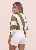 Vista posterior de blusa estampada verde oliva y blanca para mujer, con líneas anchas, mangas amplias y puños ajustados, combinada con short blanco.