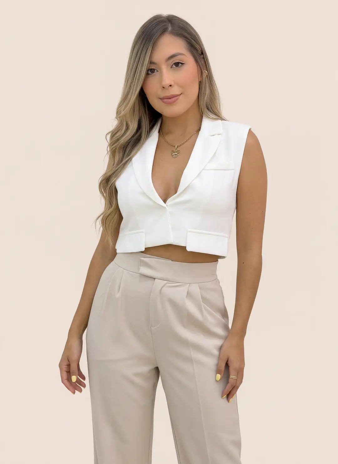 Mujer luciendo chaleco corto tipo blazer blanco sin mangas, con solapas y frente abierto, combinado con blusa blanca y pantalón beige.
