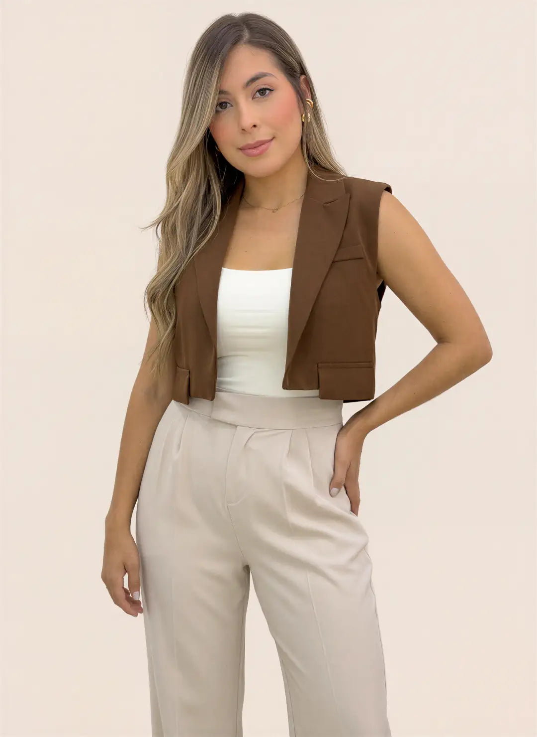 Mujer luciendo chaleco corto tipo blazer café sin mangas, con solapas y frente abierto, combinado con blusa blanca y pantalón beige.